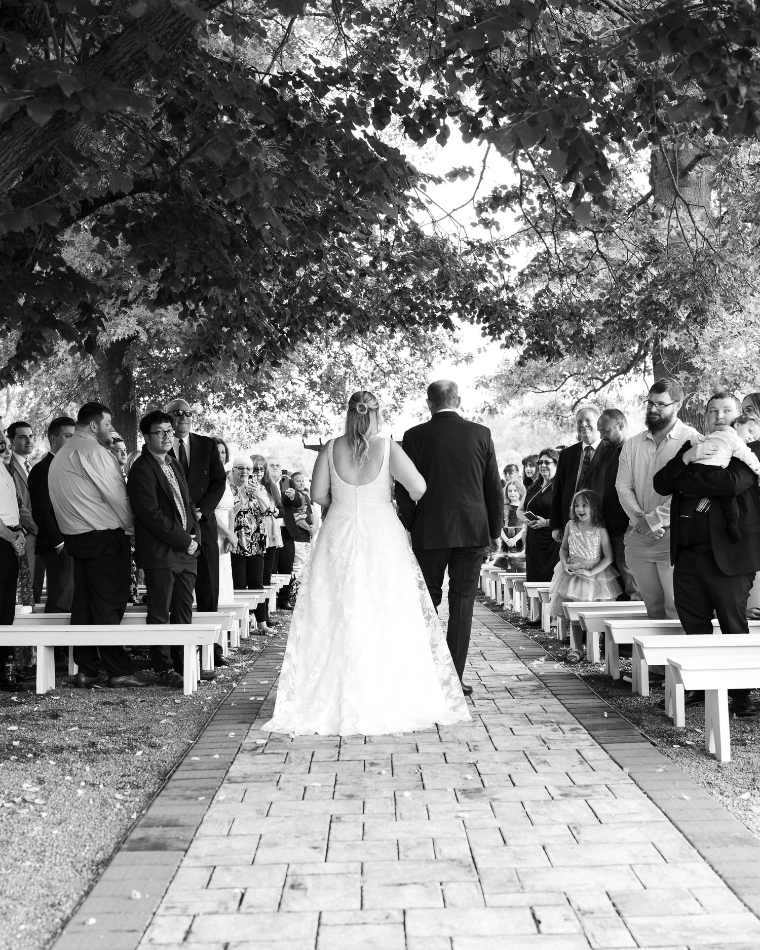 Black and white photo of bride in ball gown walking down brick aisle with escort at outdoor ceremony.