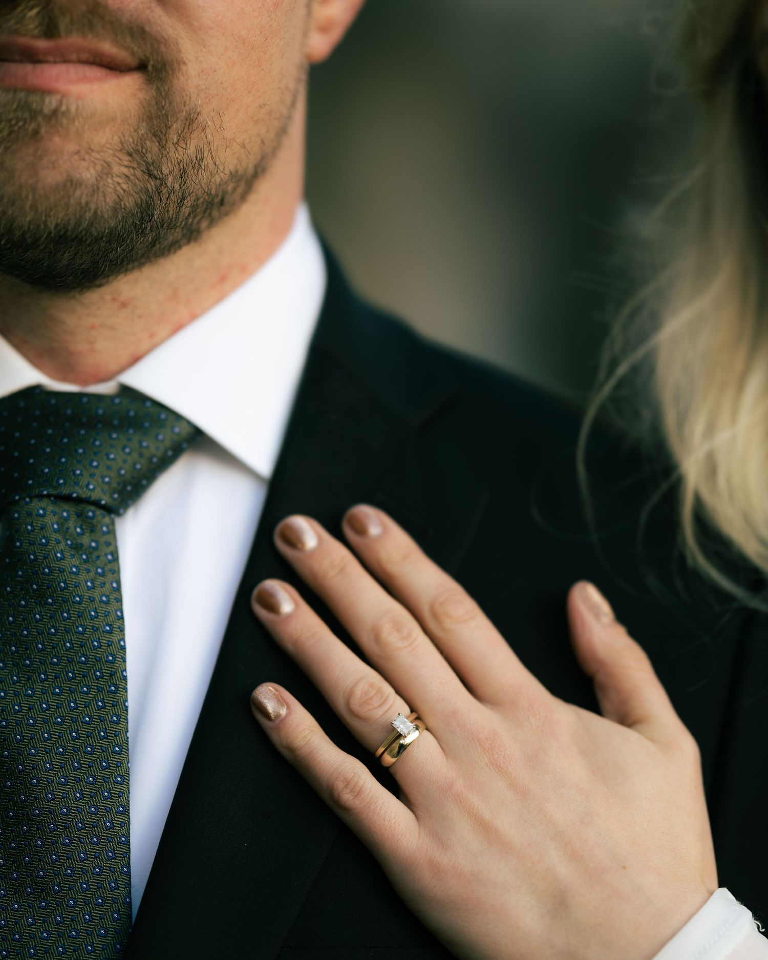 A close-up shot of a formal suit and tie with a hand wearing a wedding ring resting on the lapel.