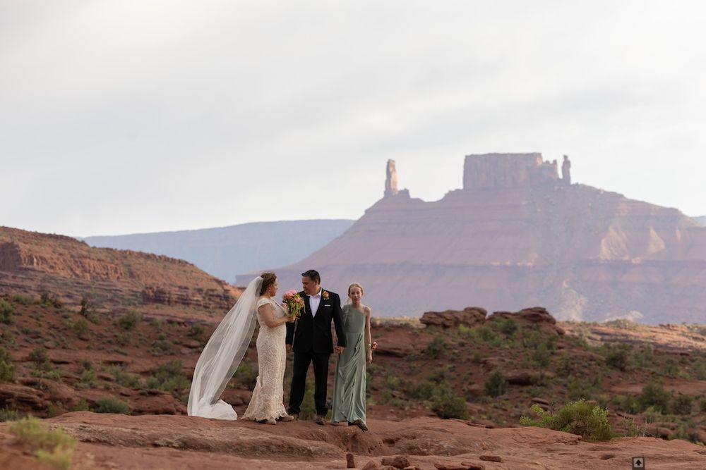 A wedding ceremony takes place on a red rock cliff with majestic desert mesas rising in the background.