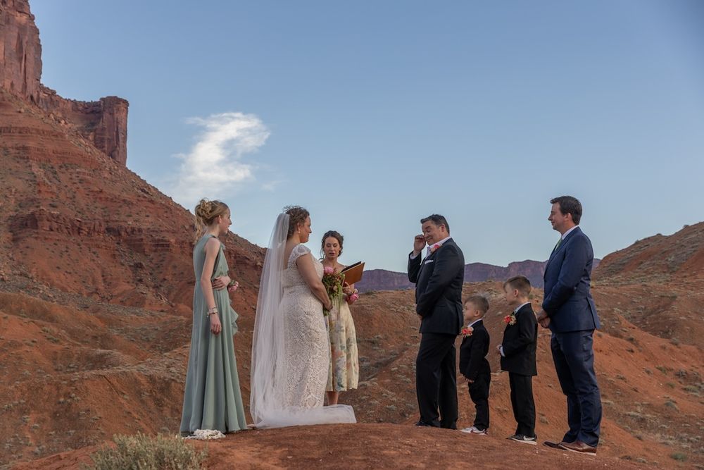 A small wedding ceremony takes place on a red rock overlook in the desert with dramatic cliffs in the background.
