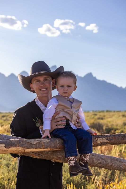 A cowboy and child sit together on a rustic wooden fence with mountain peaks and desert landscape in the background.
