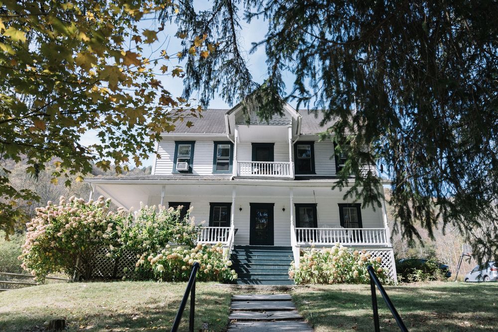 A white farmhouse with a wraparound porch sits nestled among trees and greenery in a rural setting.