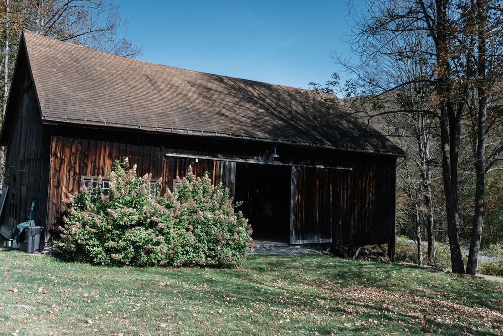 A rustic wooden barn with weathered siding stands against a backdrop of bare trees on a hillside in early spring.