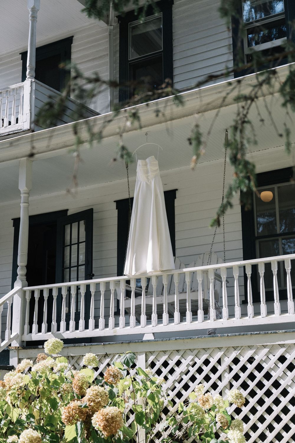 White wedding dress hanging on a porch railing with hydrangeas blooming below.