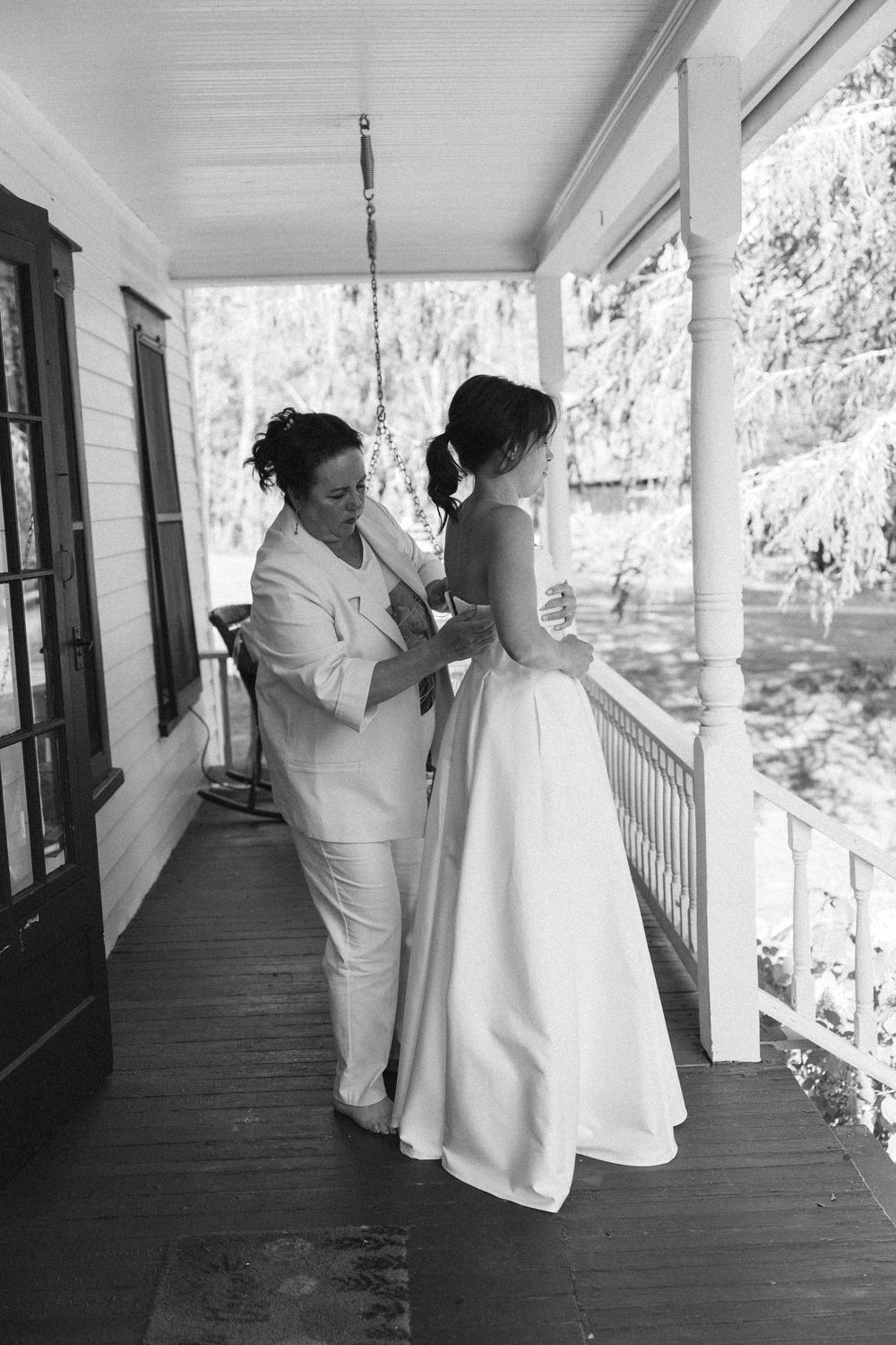 A black and white photo showing final dress adjustments on a porch of a white clapboard house.