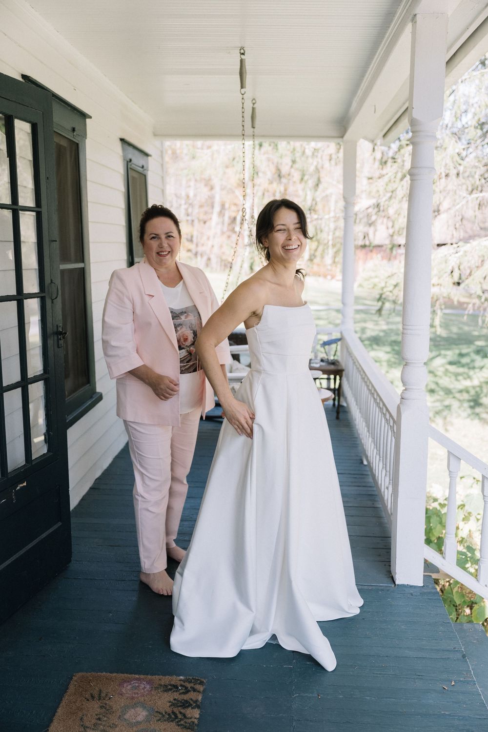 A person in a pink suit helps with a white wedding dress fitting on a covered porch.