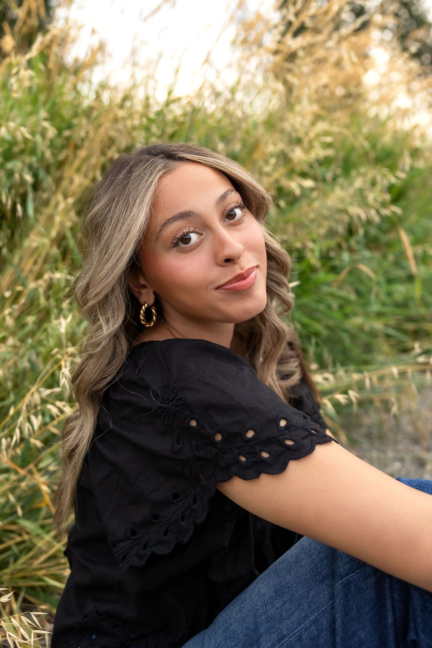 A person in a black ruffled top and jeans poses outdoors against a natural backdrop with tall grass and greenery.