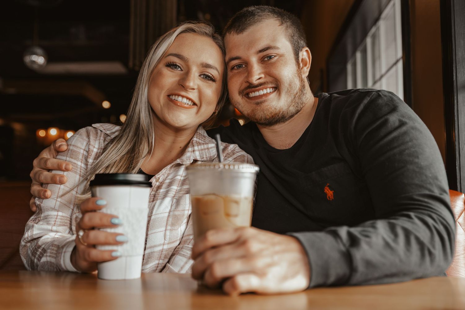 Smiling couple sitting together at a cafe table, each holding coffee drinks indoors.