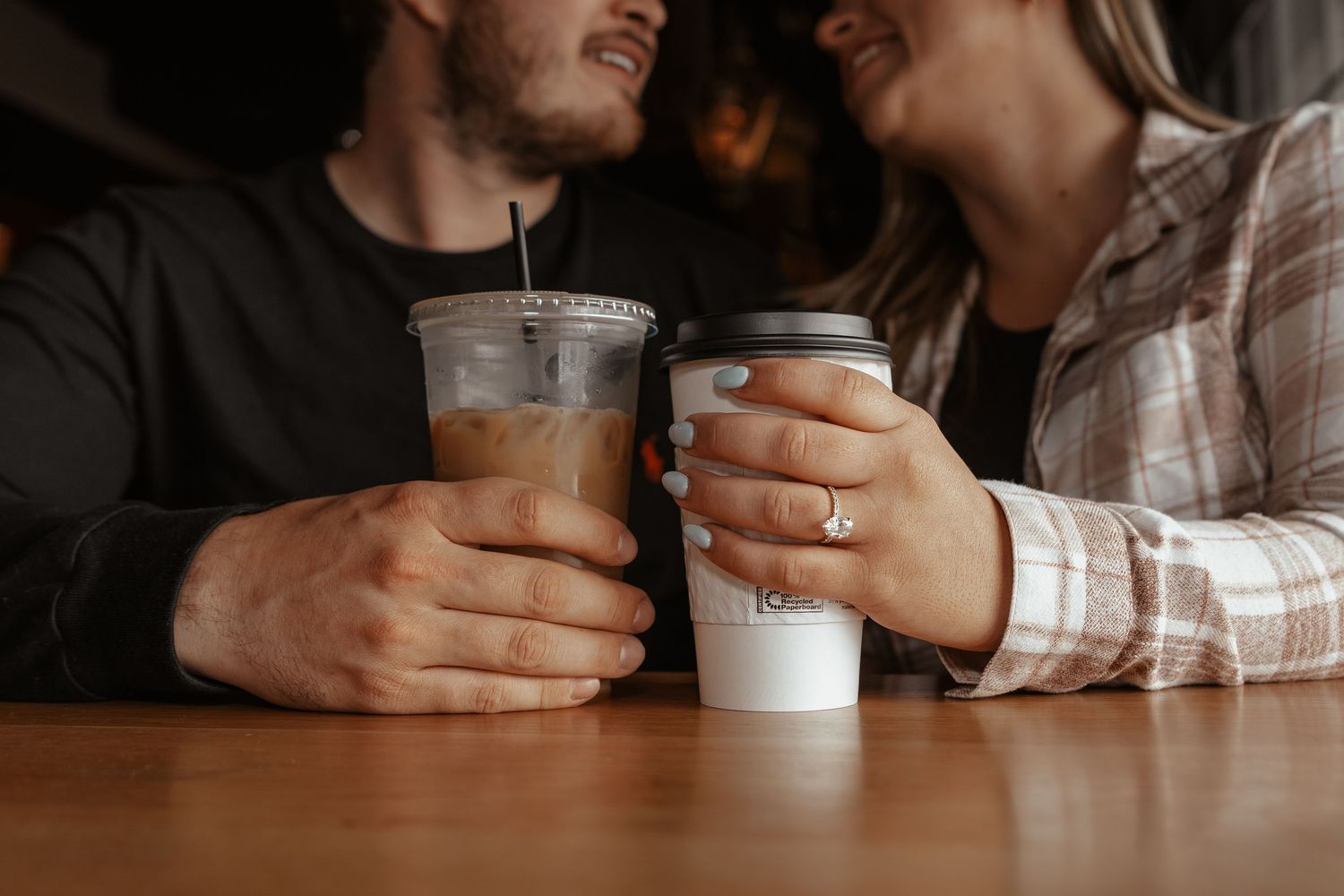 Close-up of couple holding coffee drinks, woman wearing an engagement ring, at a cafe.
