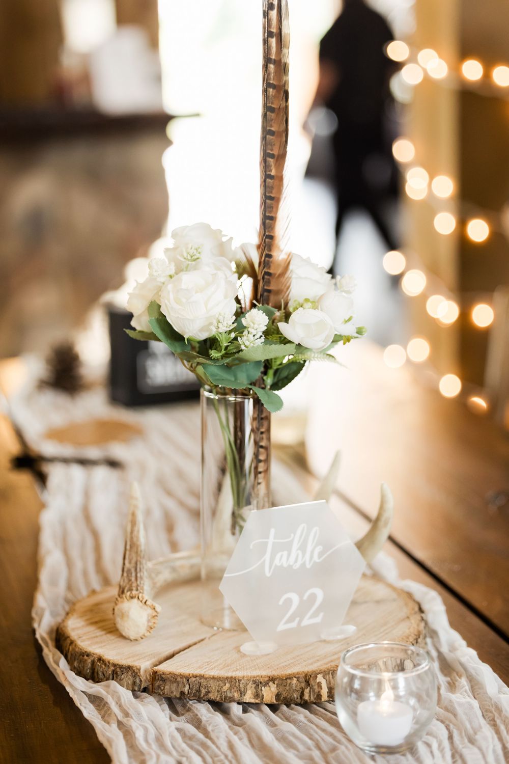 Rustic wedding table decor features white roses in glass vases on wood slices with table number cards.