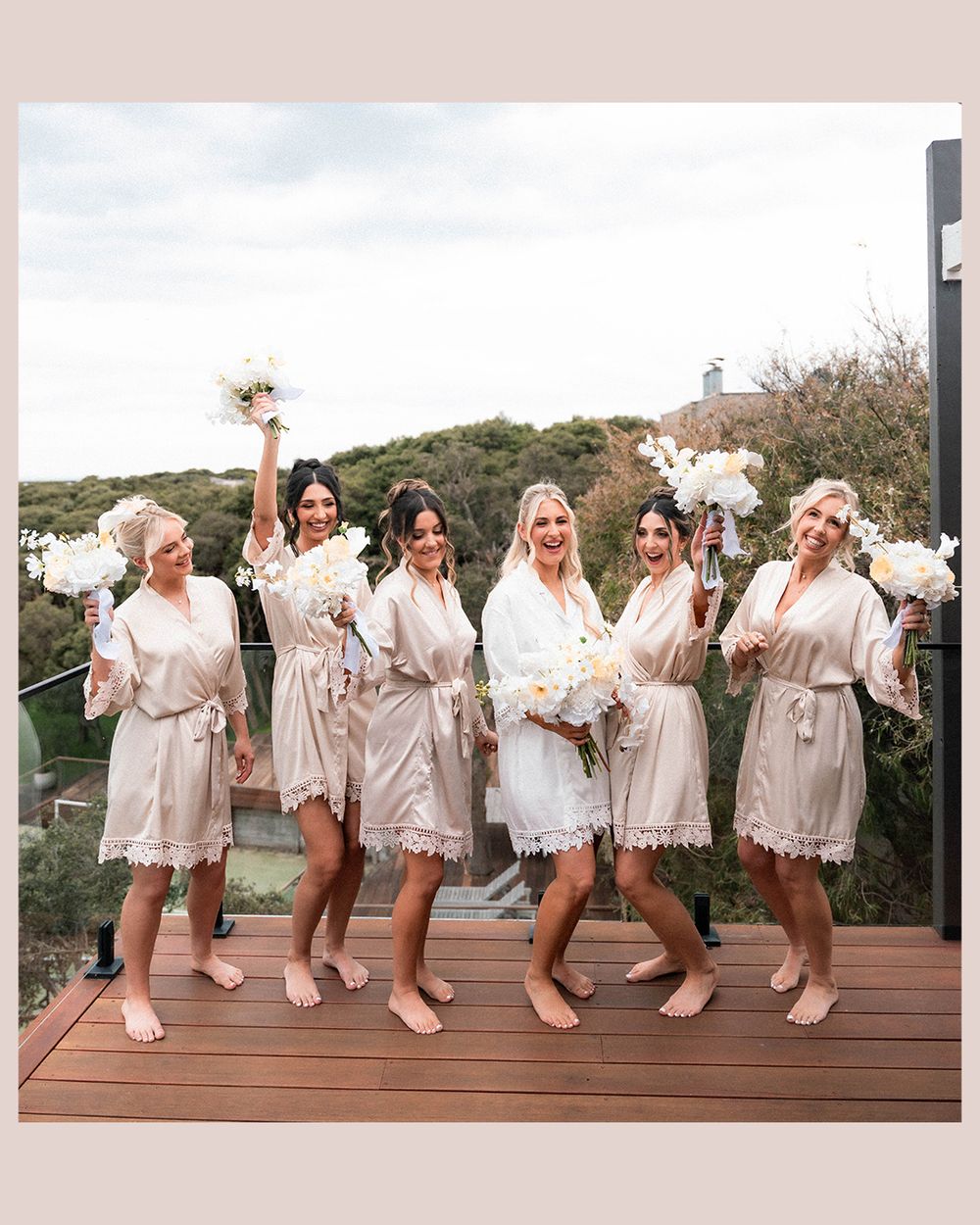 Bridal party in white robes and slippers poses playfully in a line on a wooden deck during wedding preparations.