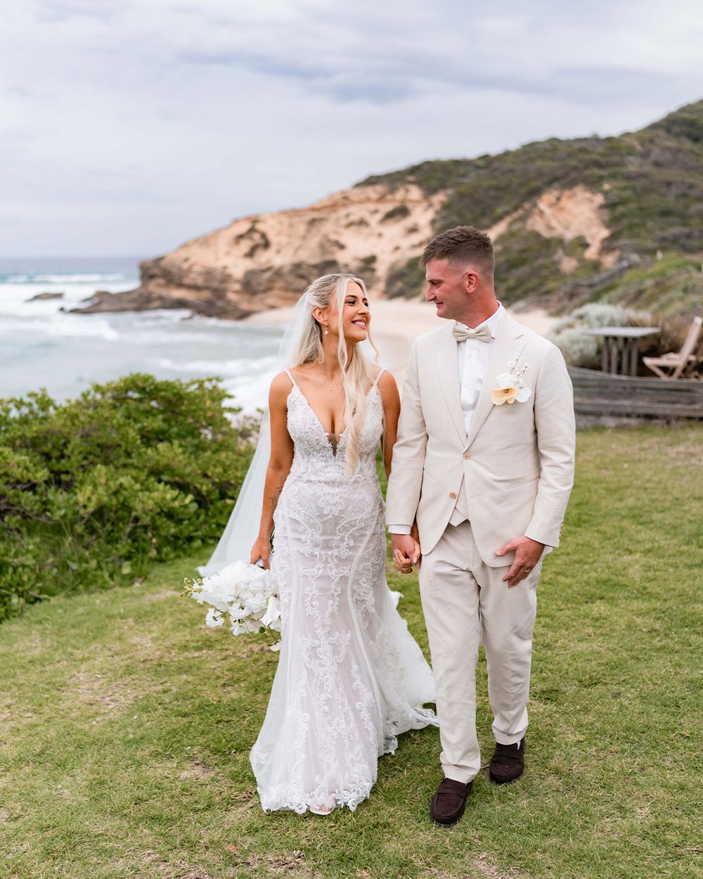 A couple in wedding attire walks together on a grassy coastal cliff with ocean views in the background.