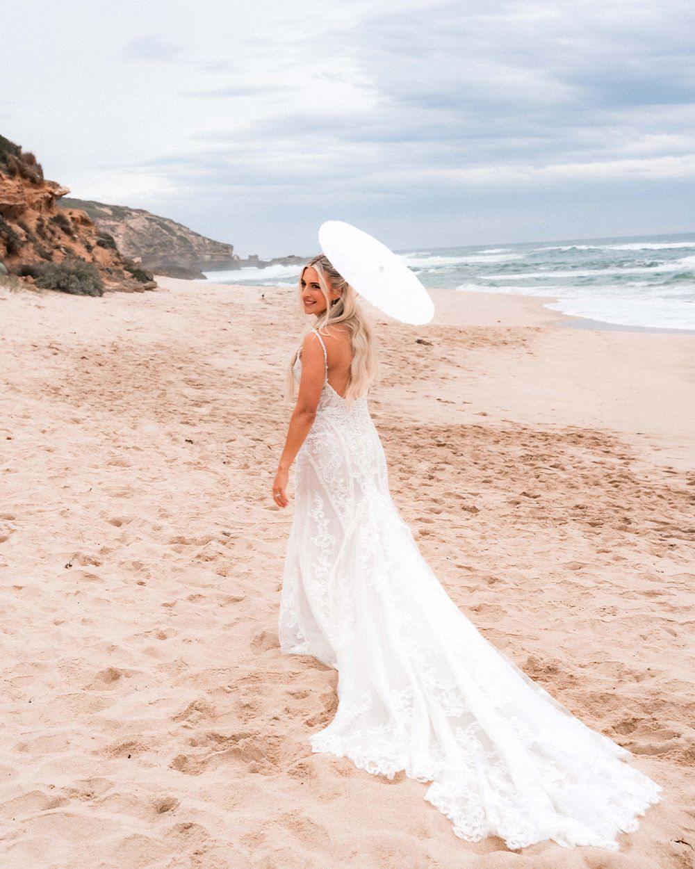 A bride in a flowing white lace gown with a long train holds a parasol while standing on a scenic beach with cliffs.