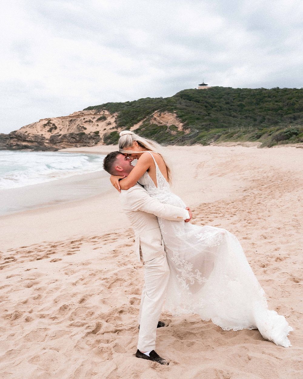 A couple in white attire playfully poses for romantic photos on a sandy beach with cliffs in the background.