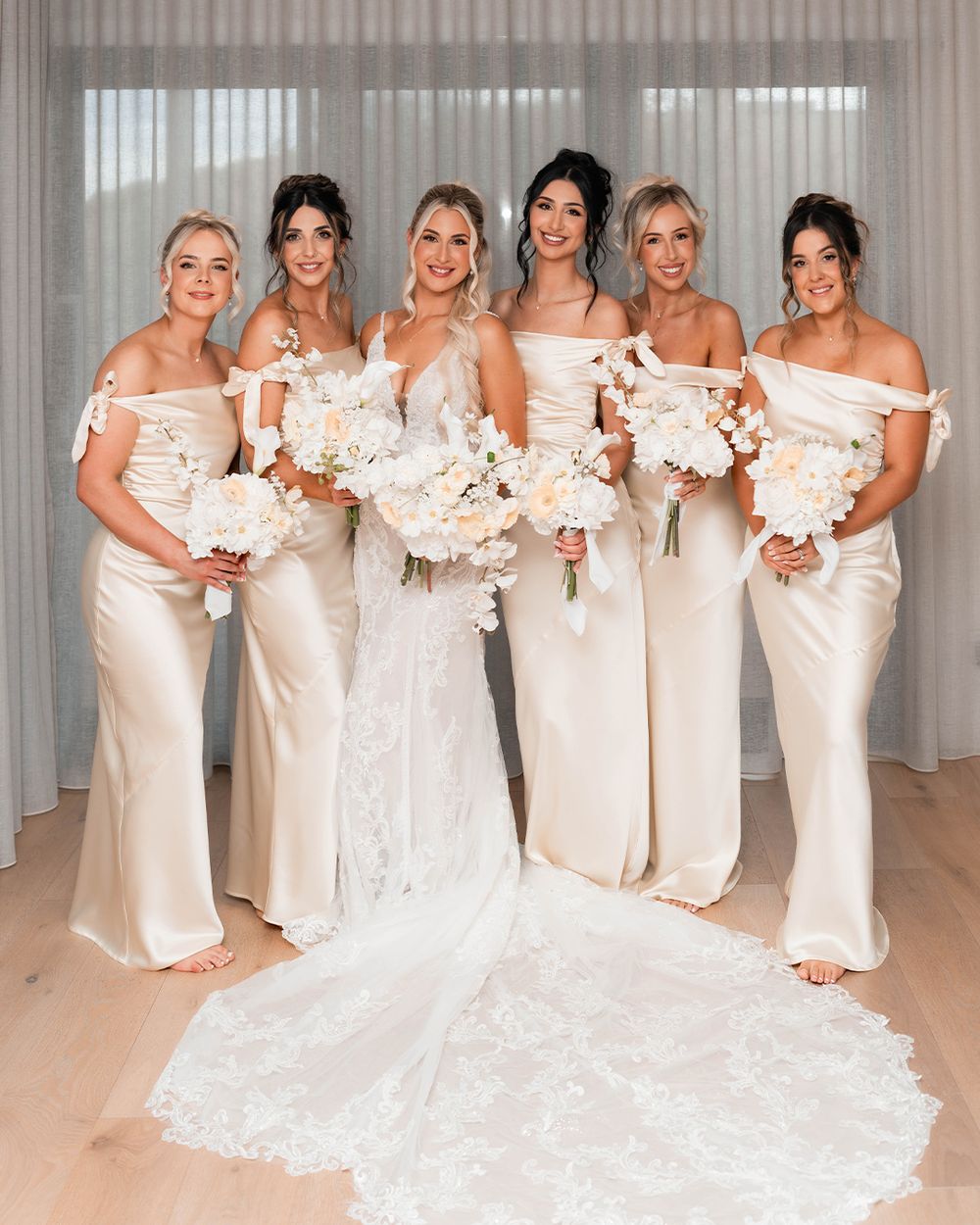 A bridal party in elegant white and cream dresses poses together holding matching floral bouquets in a sunlit room.