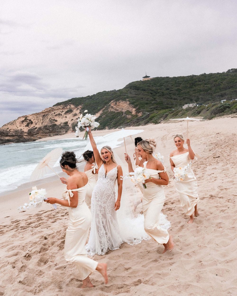 A group in white dresses playfully runs and dances along a scenic beach with waves and cliffs in the background.
