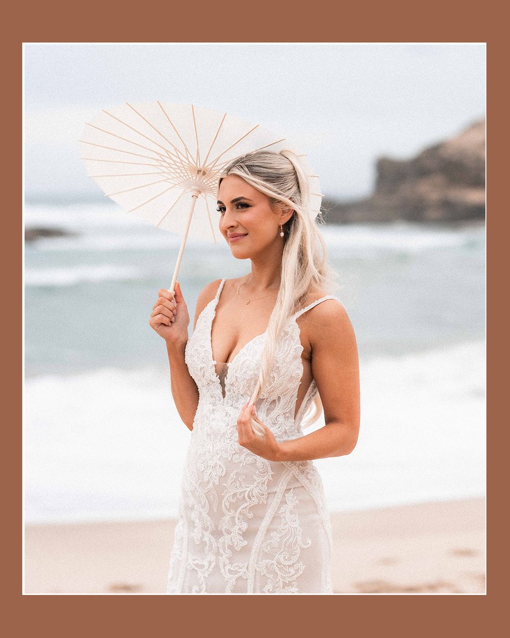 A bride in a lace wedding dress holds a peach parasol while posing on a beach with ocean waves in the background.