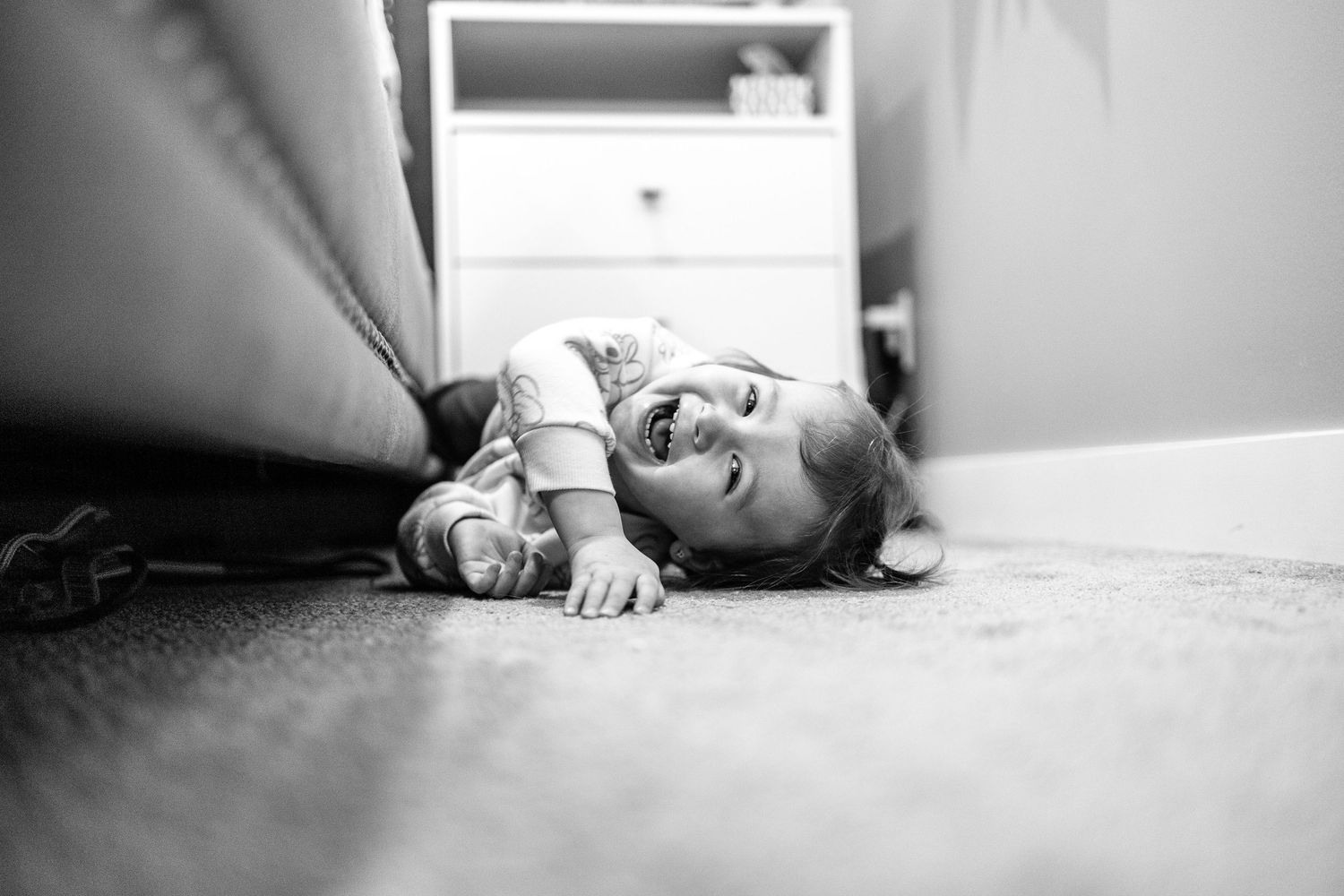 A toddler laughs while lying on the carpet next to a bed