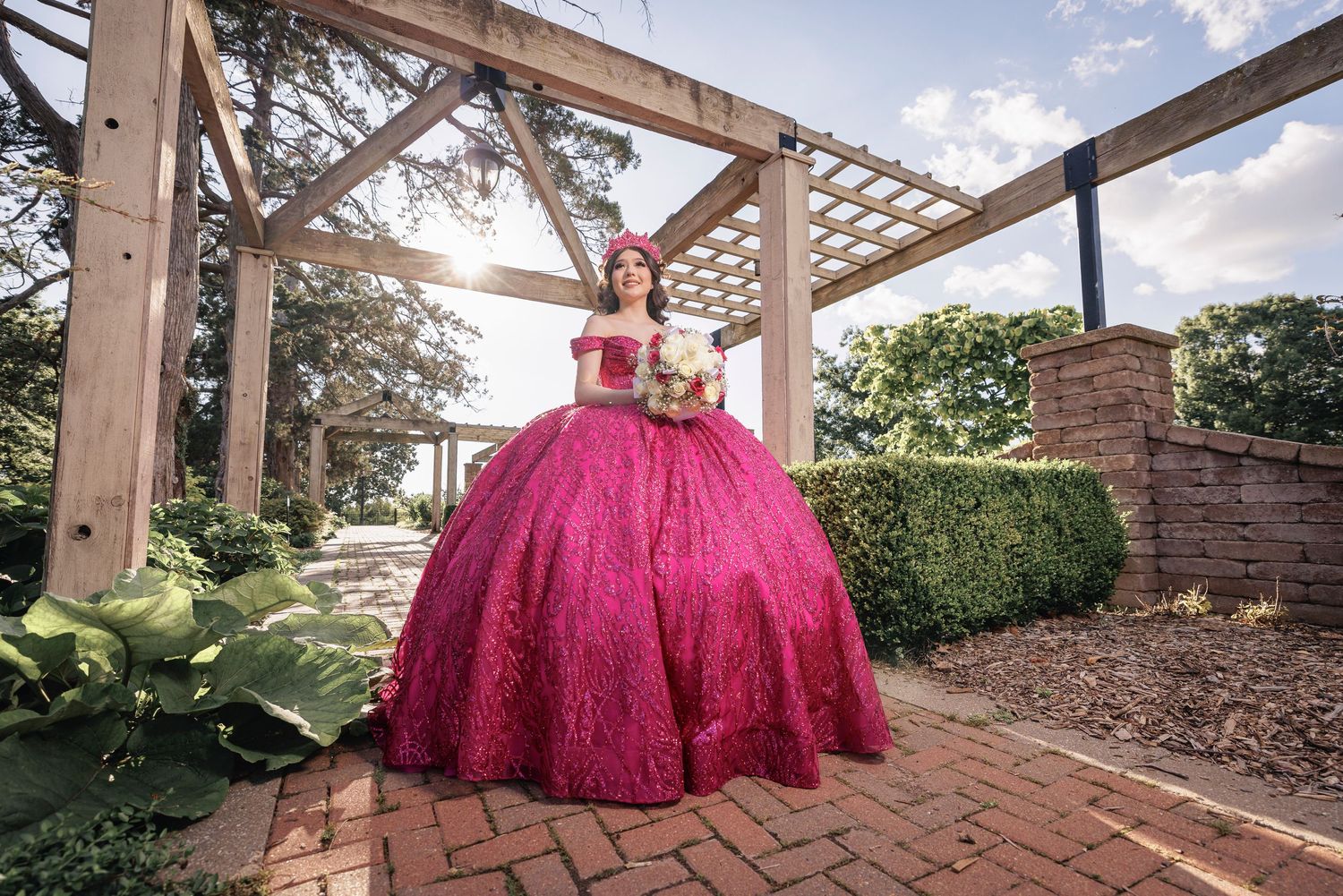 A stunning pink quinceañera ball gown showcased under a garden pergola on brick pavers.