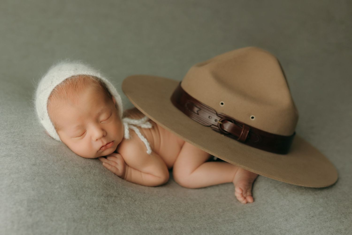 Sleeping newborn baby wearing white knit bonnet posed beside a tan park ranger hat on a gray background at the newborn photo studio.
