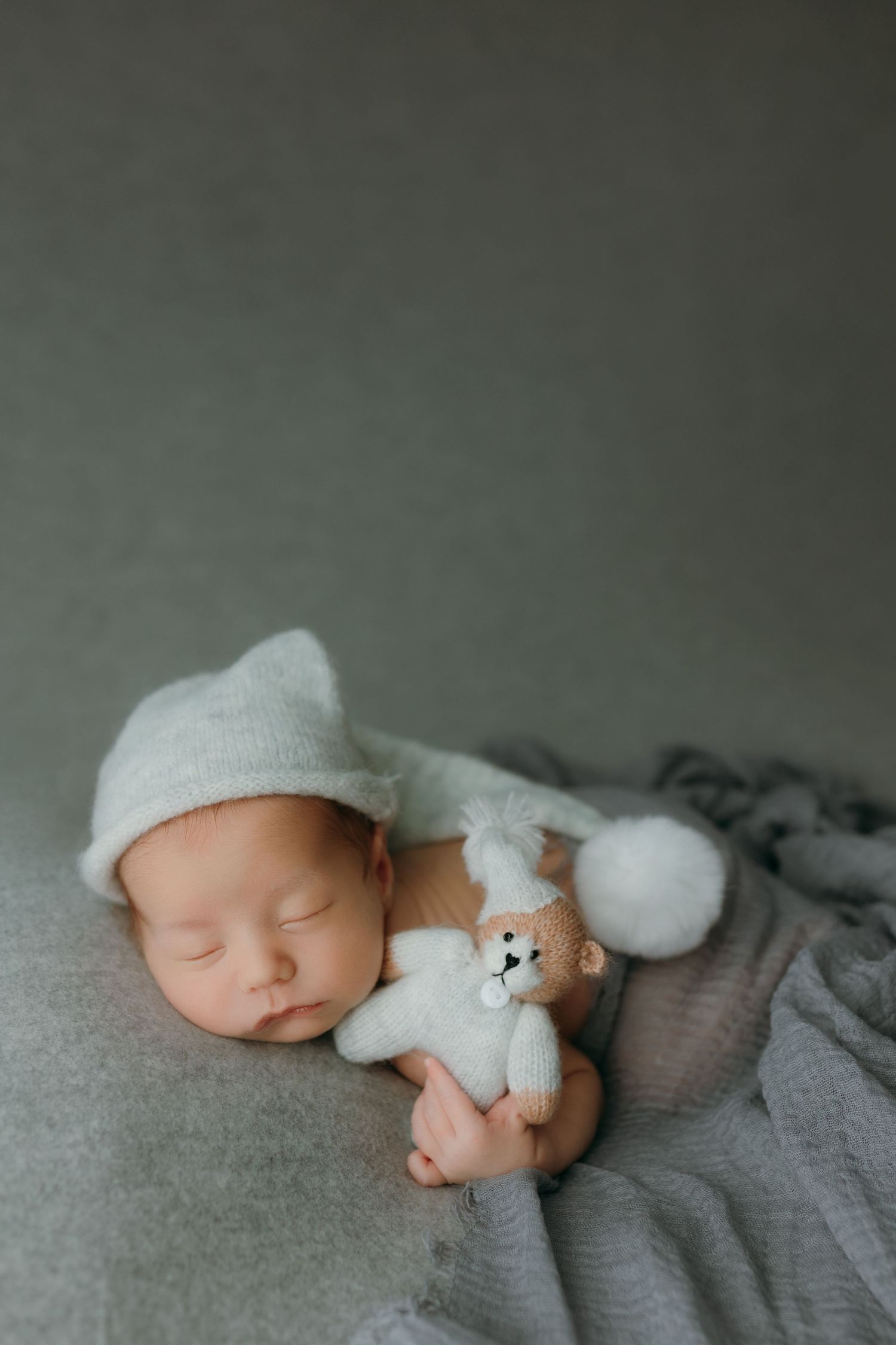 Sleeping newborn baby wearing a white knit hat holding a tiny teddy bear on a soft gray blanket at the newborn photo studio in victoria bc.