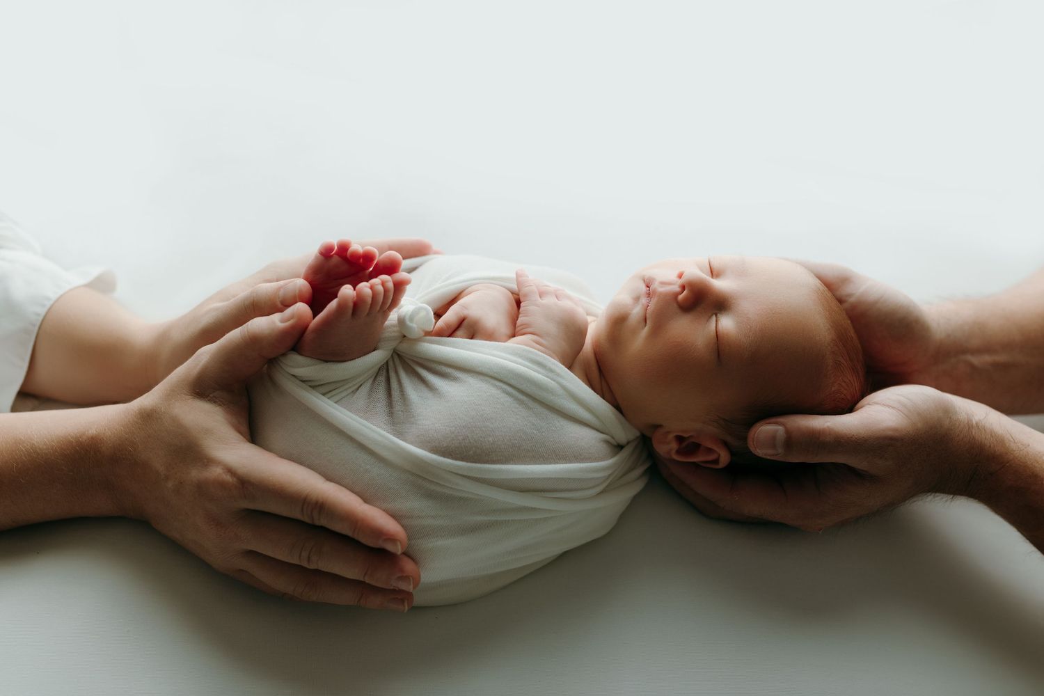Newborn baby swaddled in white wrap, cradled gently by parents hands on a soft white background at the newborn photo studio in victoria bc.