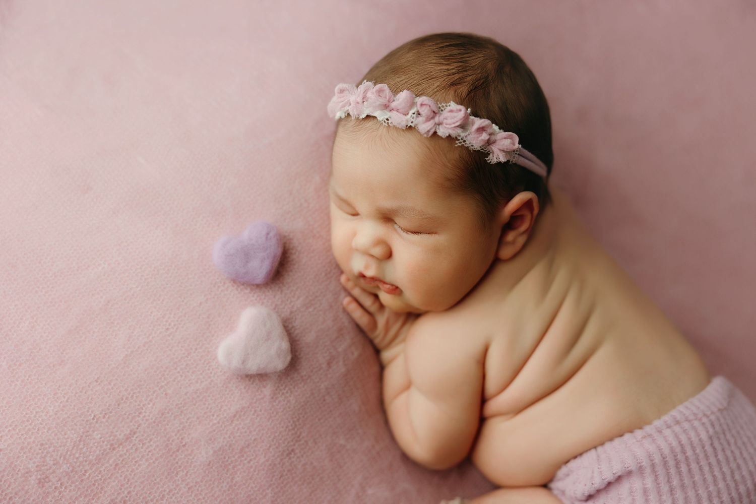 Sleeping newborn baby girl wearing a pink floral headband, posed on a pink blanket with soft heart-shaped props at newborn photo studio