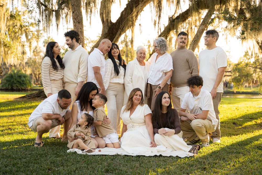 Large family portrait outdoors under Spanish moss trees, wearing neutral tones of white and beige, gathered on green grass.