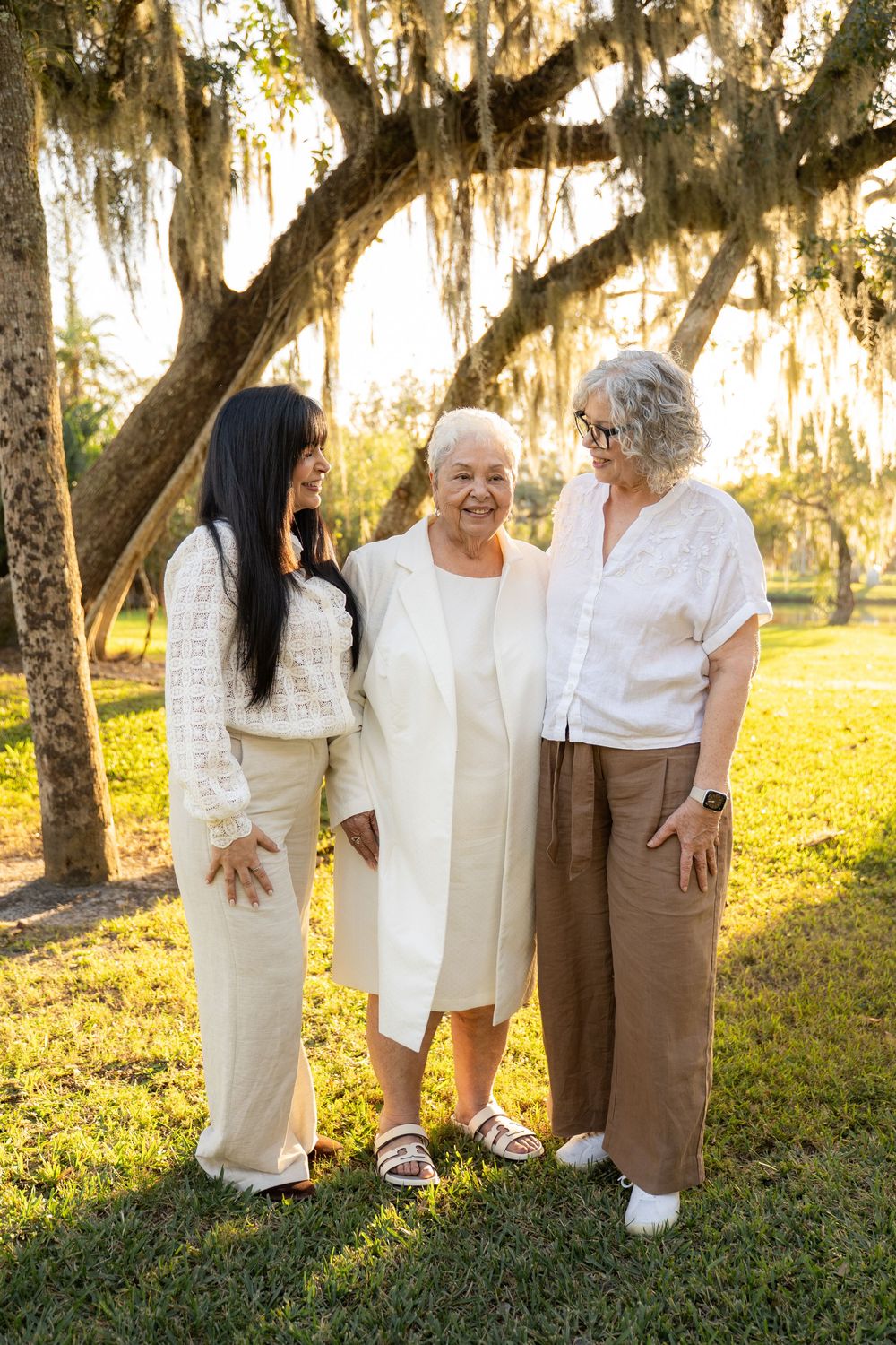 Three women smiling together outdoors under Spanish moss-draped trees in warm golden sunlight.