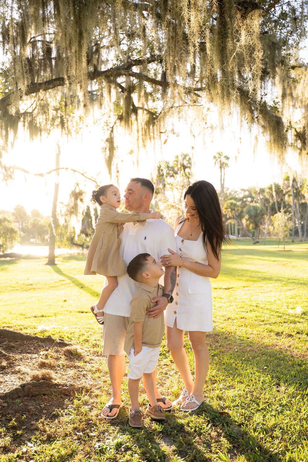 Family of four posing under Spanish moss-draped oak trees in a sunlit park, wearing coordinated neutral outfits.
