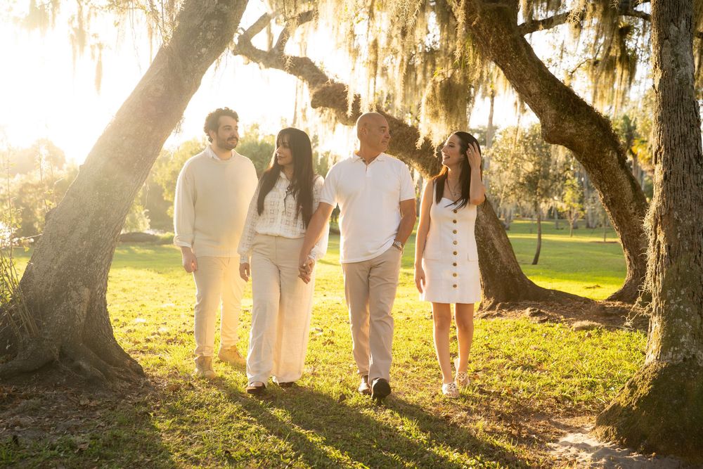 Family of four walking under moss-draped oak trees in golden sunlight, dressed in white outfits on lush green grass.