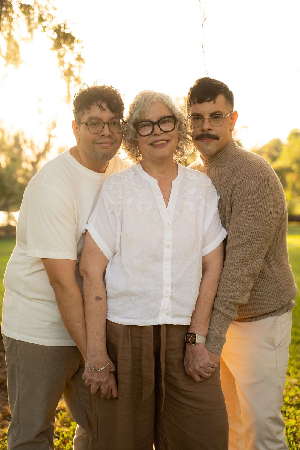 Three smiling family members pose together outdoors during golden hour sunset in a park setting.