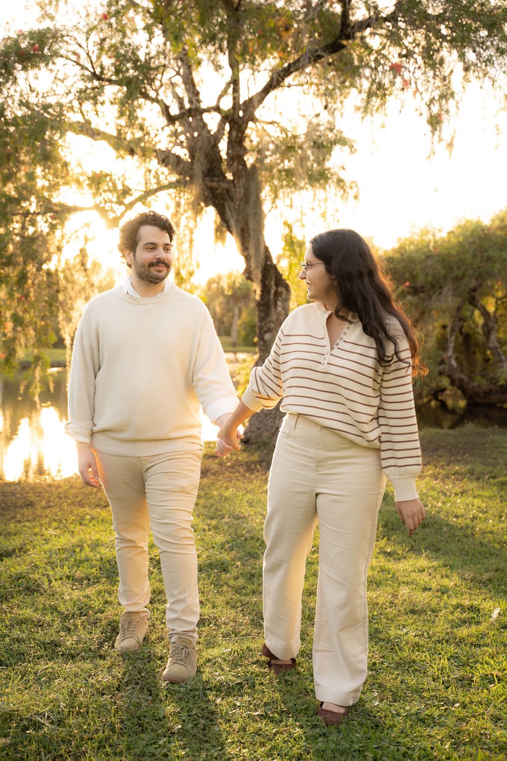 Couple walking hand in hand outdoors at golden hour, wearing matching cream outfits near a serene pond with trees.