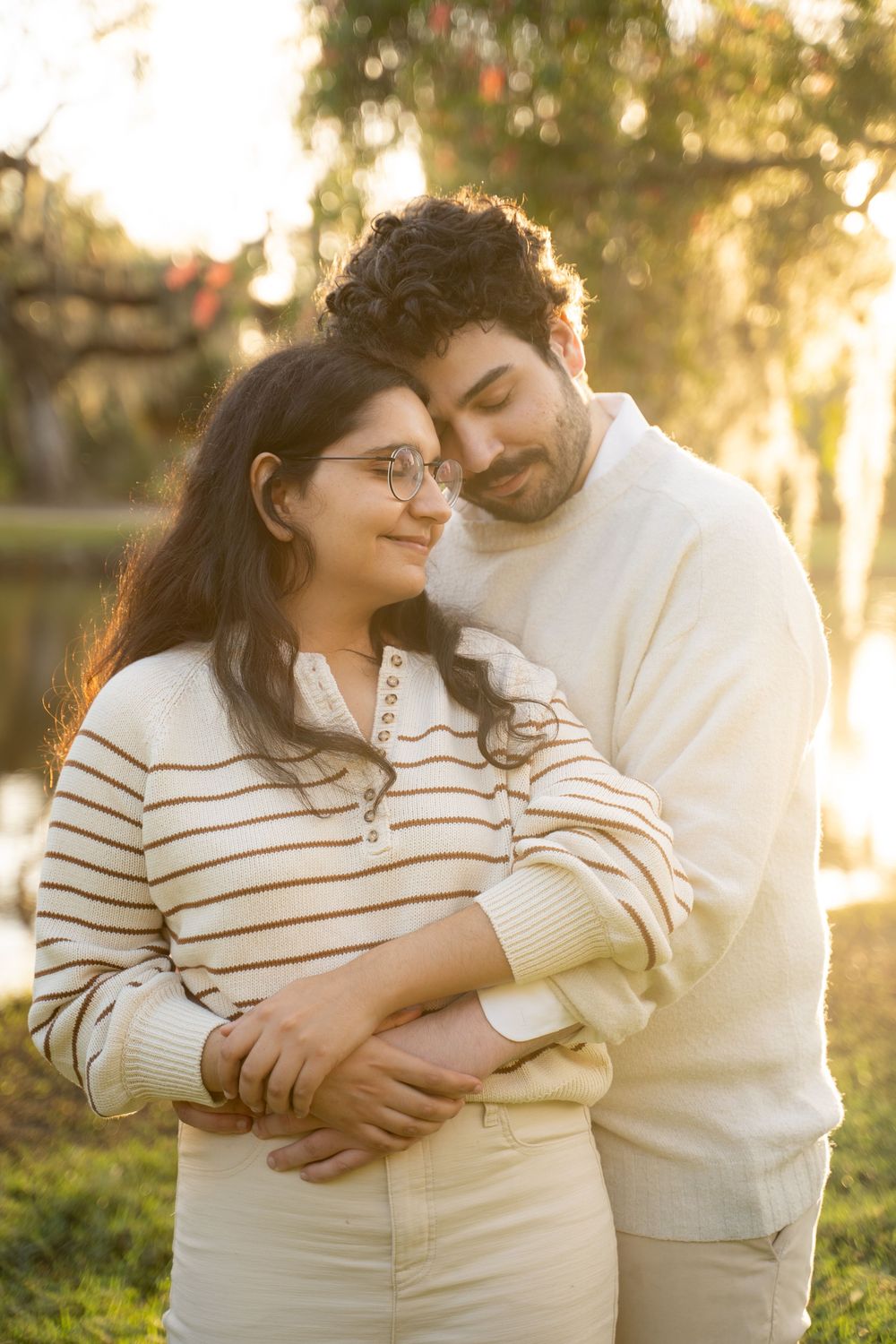 Couple embracing outdoors at golden hour, woman in striped sweater with glasses, man in cream sweater, soft bokeh background.