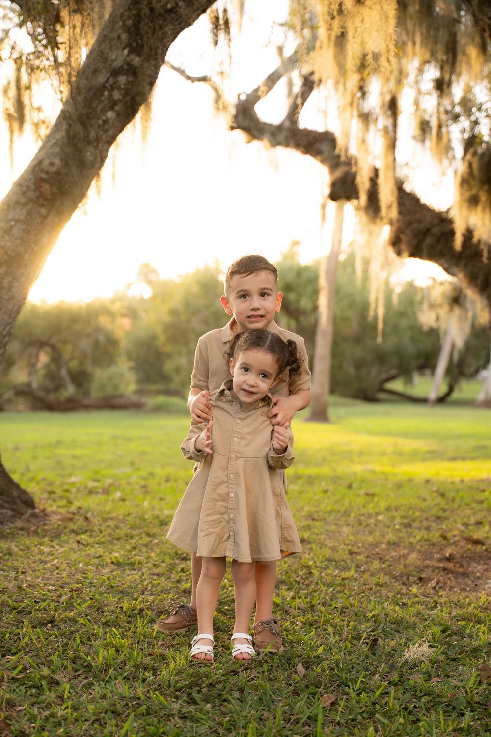 Two children in matching khaki outfits embrace under a moss-draped oak tree at golden hour in a lush green park.