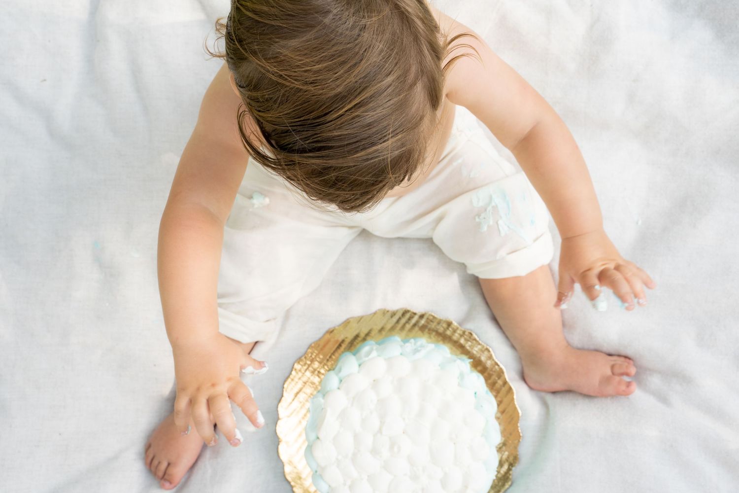 Overhead view of a baby in white outfit reaching into a small cake with white frosting on a gold plate.
