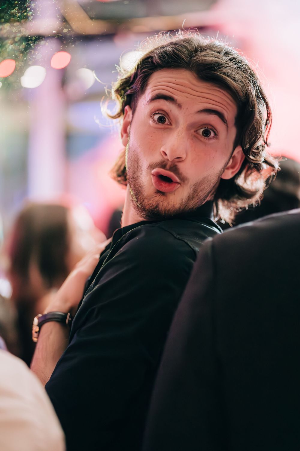 Surprised young man with curly hair and beard at a social event, wearing black shirt.