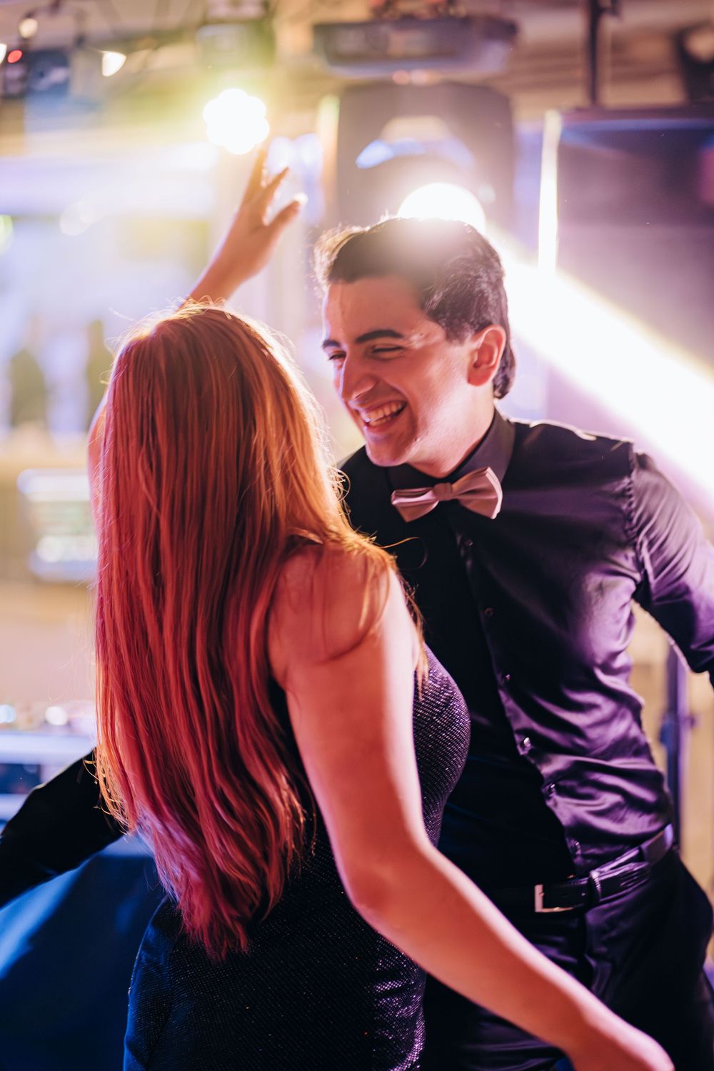 A smiling man in a black shirt and bow tie dances with a red-haired woman in a sparkly dress at a nightclub.