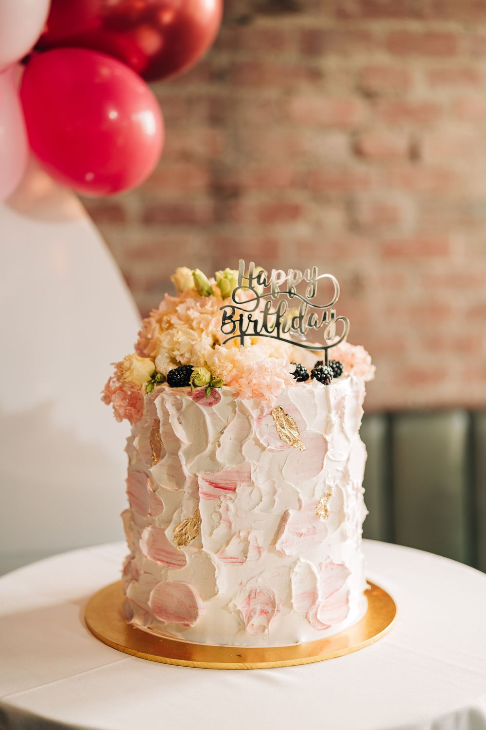 Elegant white birthday cake with textured frosting, pink flowers, and a gold Happy Birthday topper on a white table.