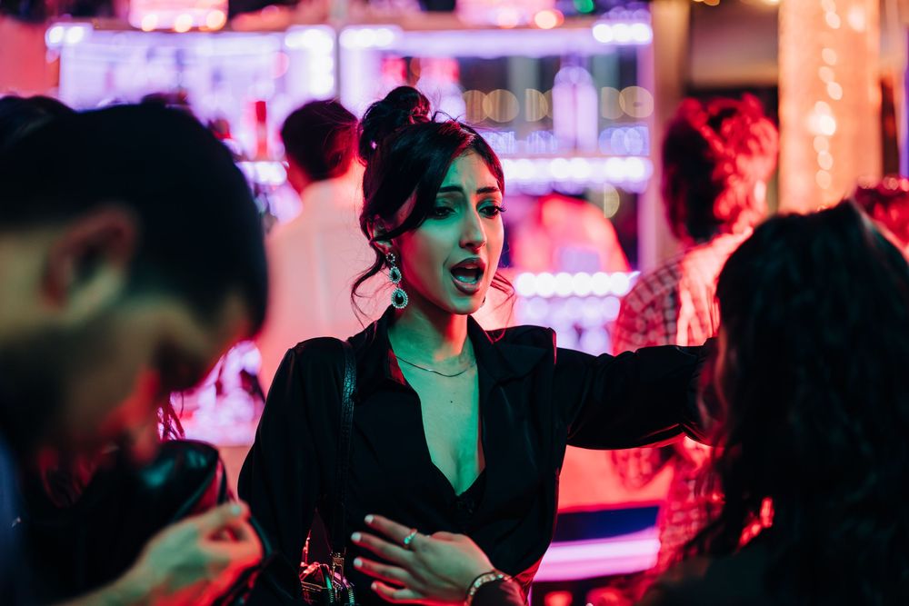 Woman in black blazer socializing at a vibrant neon-lit bar or nightclub party.