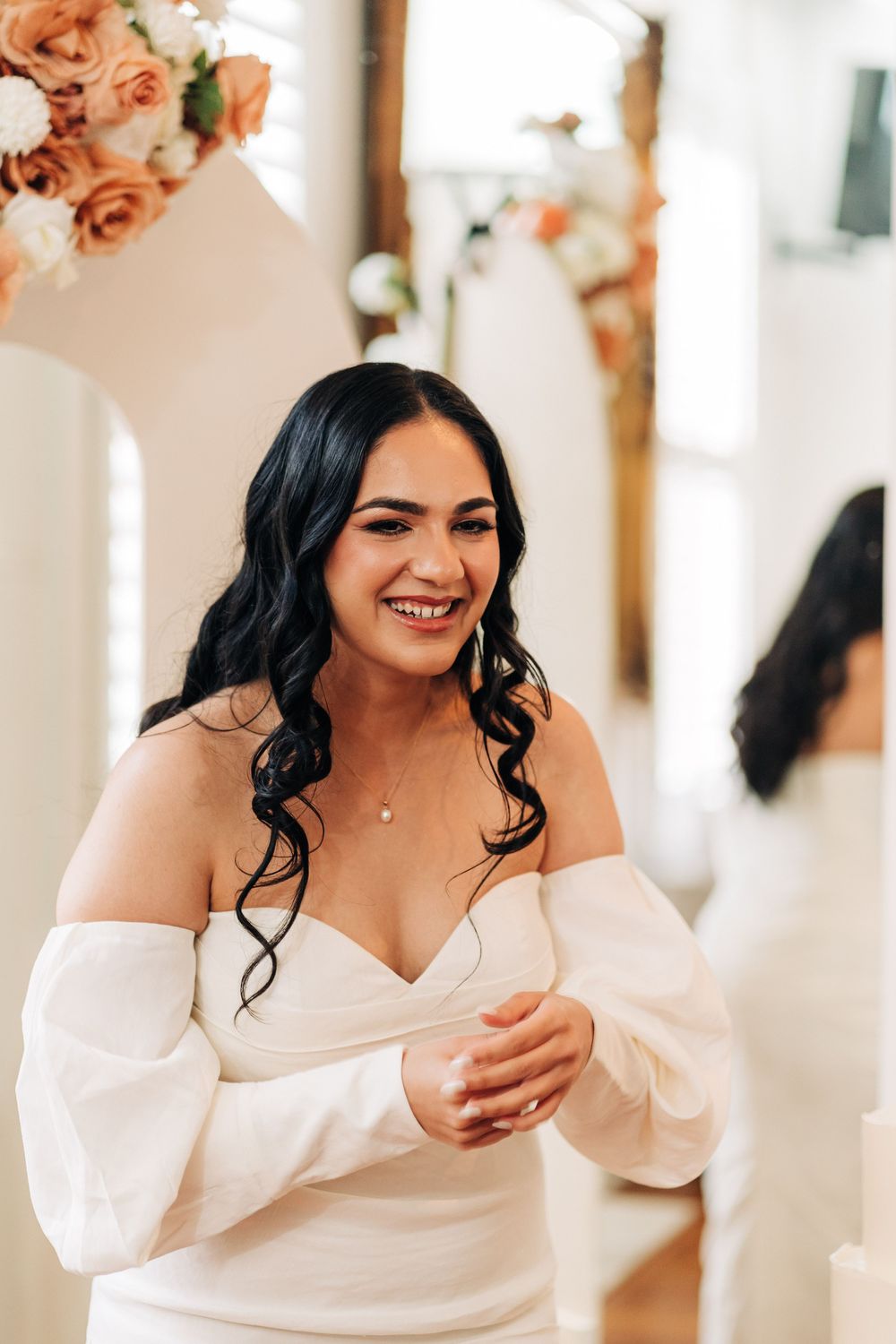 Smiling bride in off-shoulder white gown with dark wavy hair, standing near floral arrangements in bright venue.
