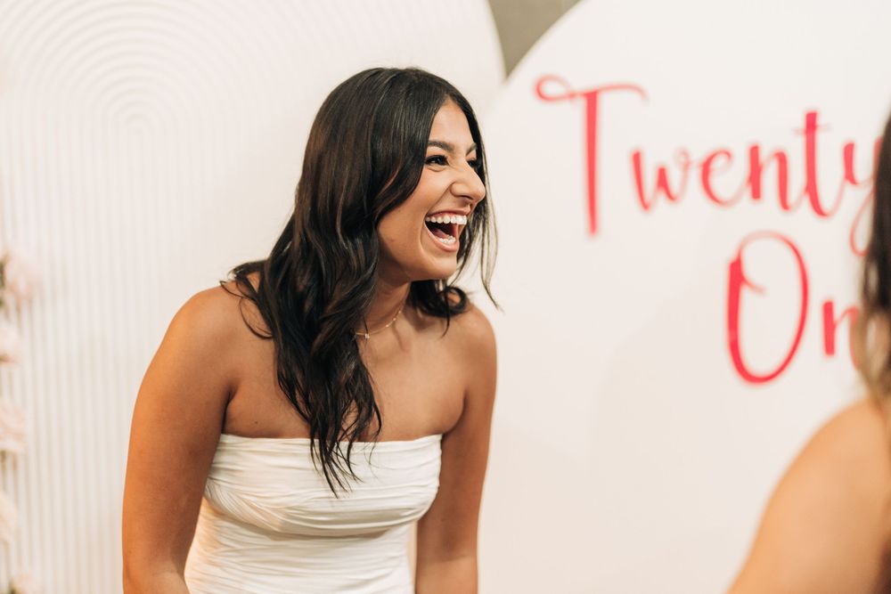 A smiling woman in a white strapless dress laughs joyfully at a Twenty One themed event.