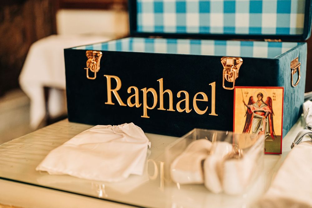 Personalized navy blue trunk with gold "Raphael" lettering beside a religious icon and white baptism outfit.