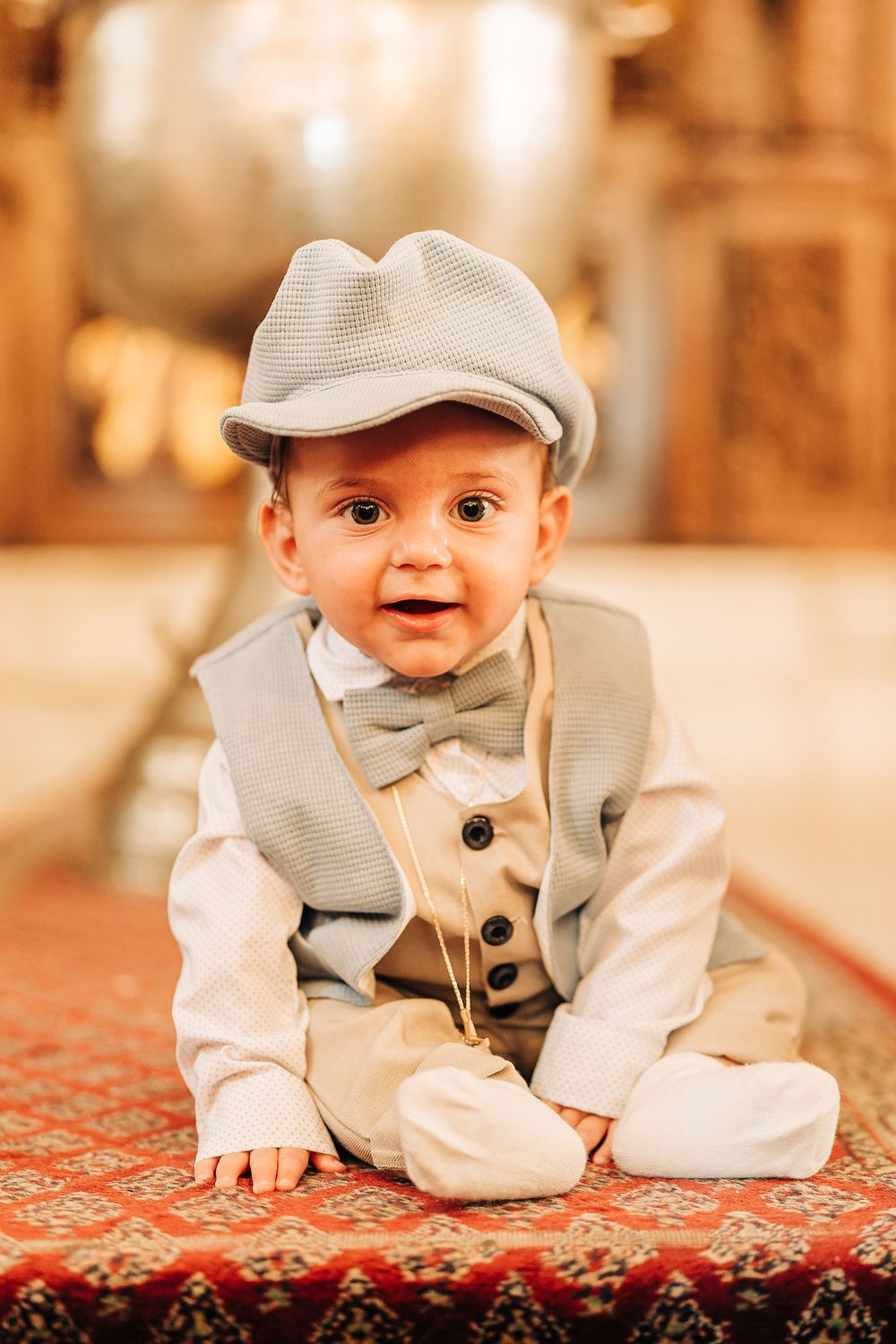 Adorable baby boy in vintage gray vest, flat cap, and bow tie sitting on a red patterned rug.
