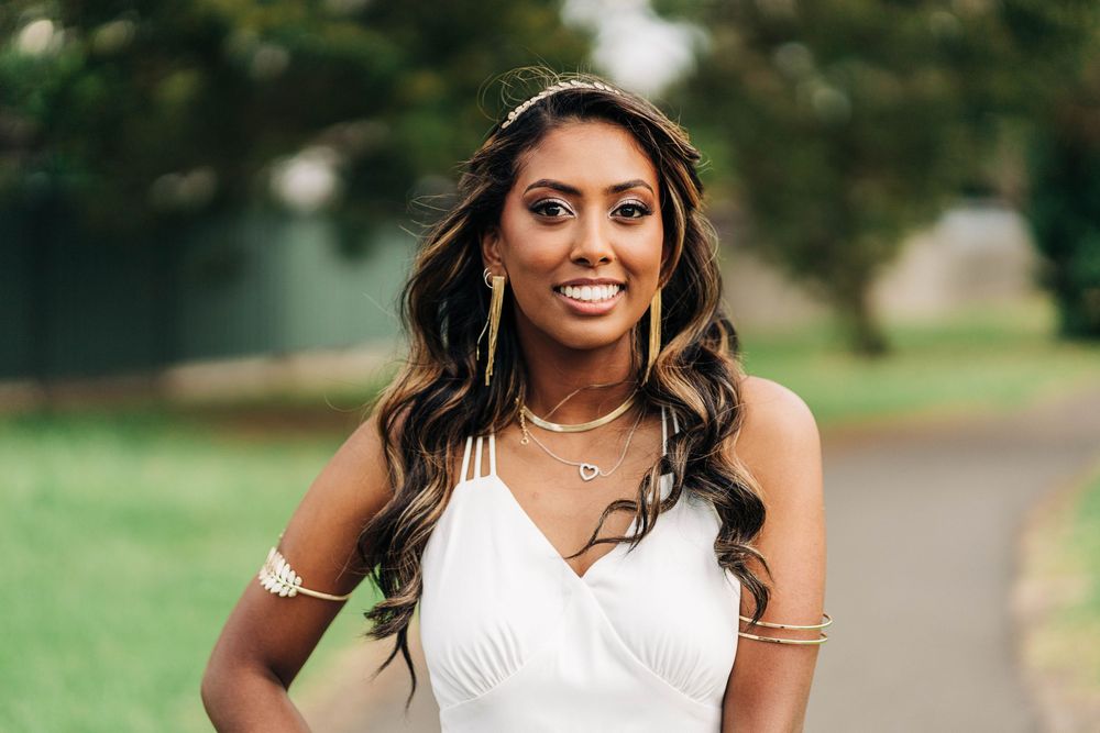 Woman in white dress with gold jewelry smiling outdoors in a park setting.