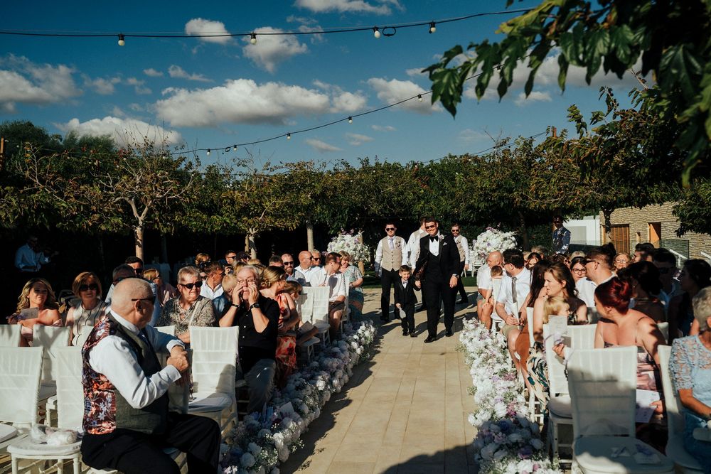 Outdoor wedding ceremony with guests seated on white chairs under a bright blue sky with string lights overhead.