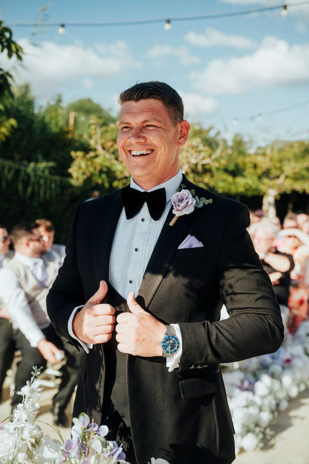 A person in a black tuxedo and bowtie smiles and gestures during an outdoor celebration with string lights visible.