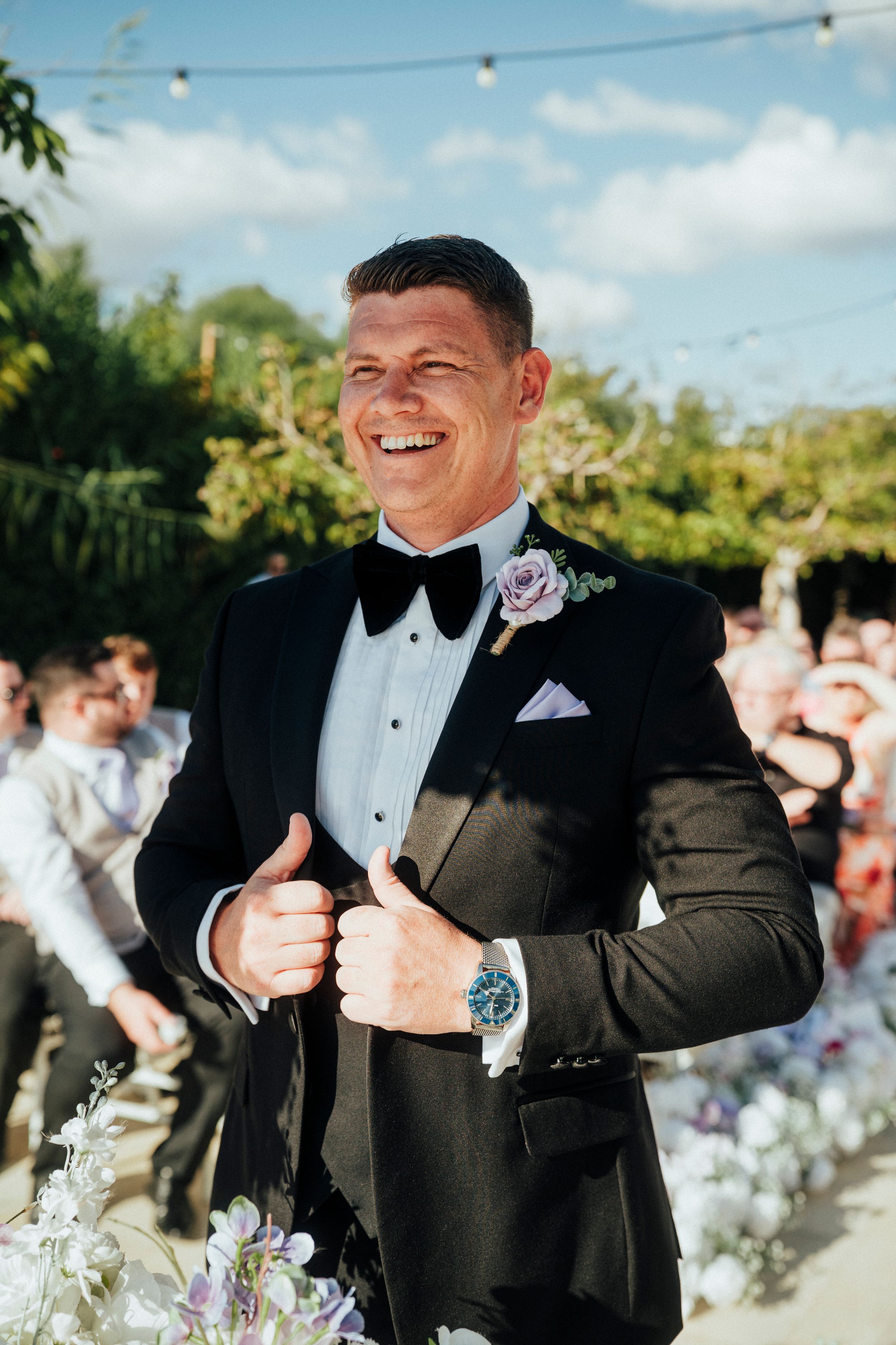 A person in a black tuxedo and bowtie smiles and gestures during an outdoor celebration with string lights visible.