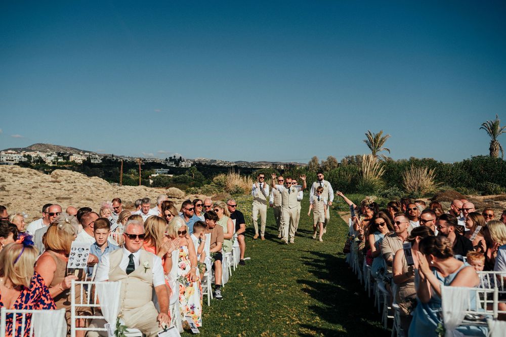 Outdoor wedding ceremony on a grassy hilltop with guests seated in white chairs under blue sky and palm trees.