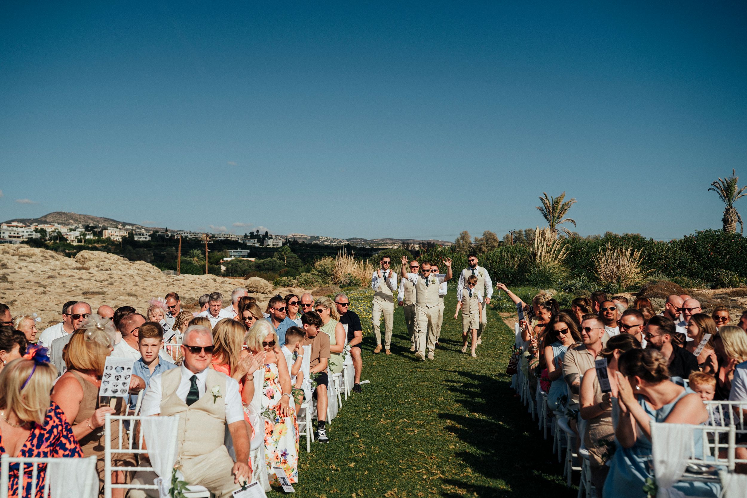 Outdoor wedding ceremony on a grassy hilltop with guests seated in white chairs under blue sky and palm trees.
